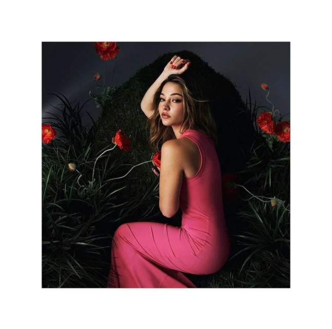 Portrait of a woman in a pink dress reclining among long grasses and red flowers, captured in a softly lit, intimate setting.