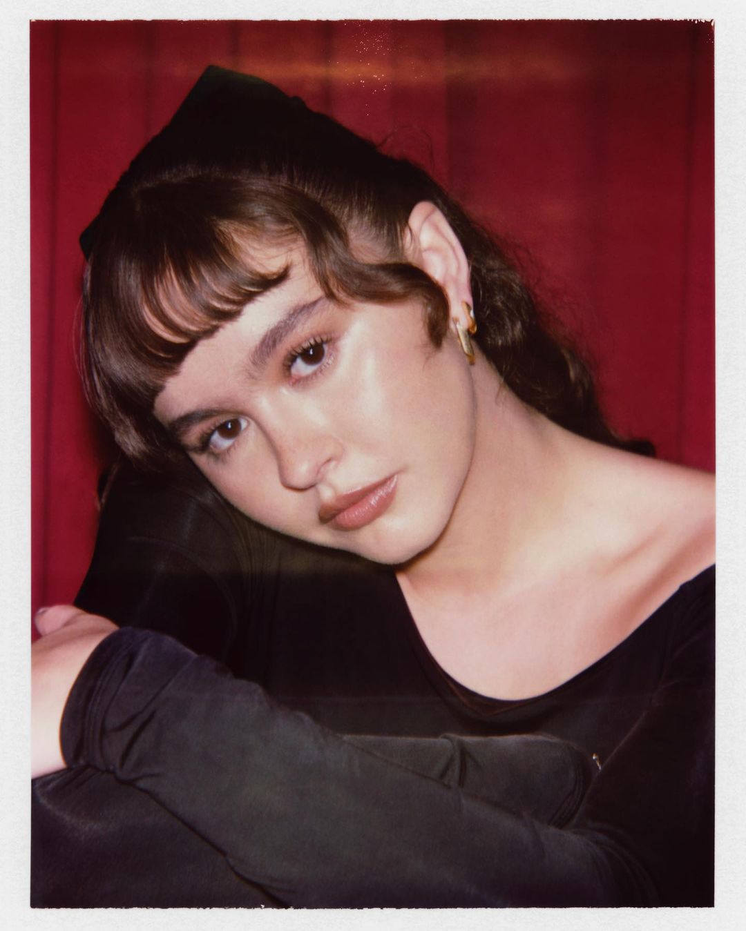 Polaroid portrait of a woman in a black off-the-shoulder top against a pleated red curtain background.
