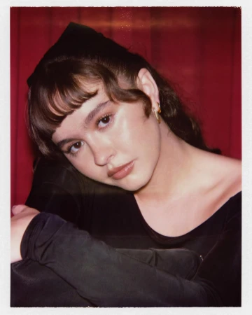 Polaroid portrait of a woman in a black off-the-shoulder top against a pleated red curtain background.