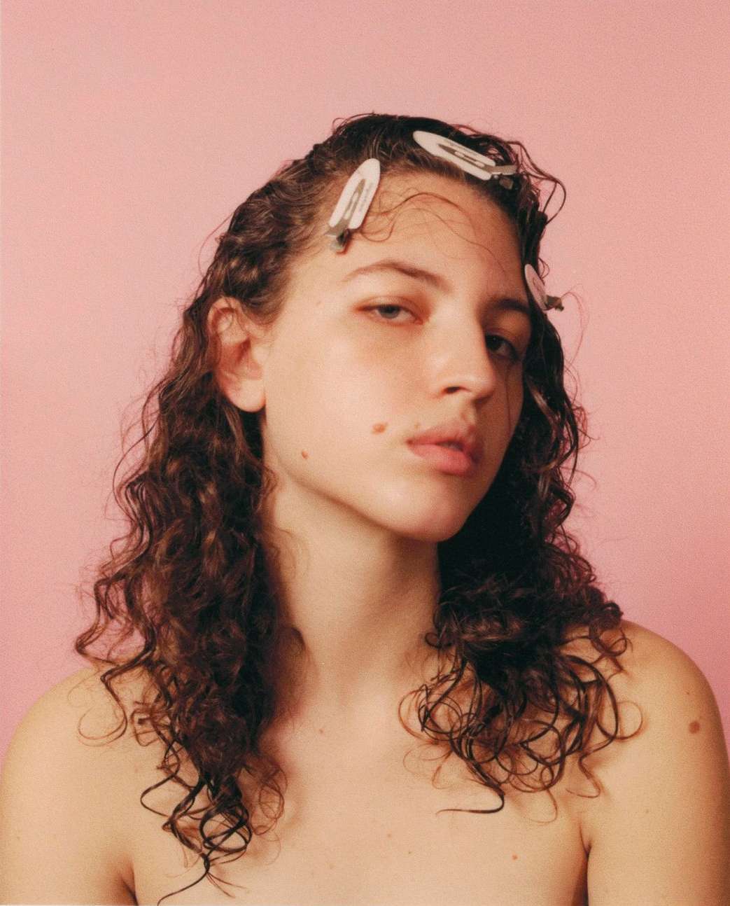 Close-up portrait of a young adult woman with curly brown hair, several white hair clips, light skin, and a pink background.