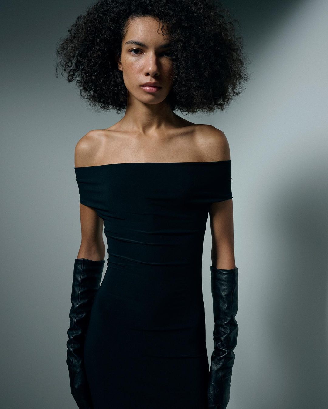 Woman with curly hair poses in a black, off-the-shoulder dress and long gloves in a photo studio setting with soft, directional lighting.