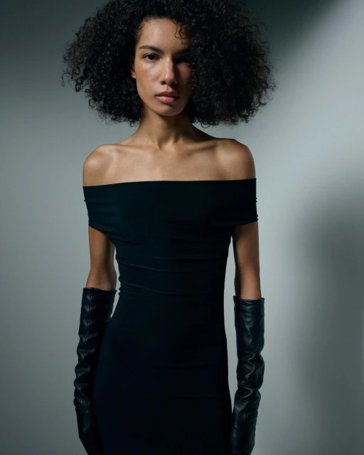 Woman with curly hair poses in a black, off-the-shoulder dress and long gloves in a photo studio setting with soft, directional lighting.