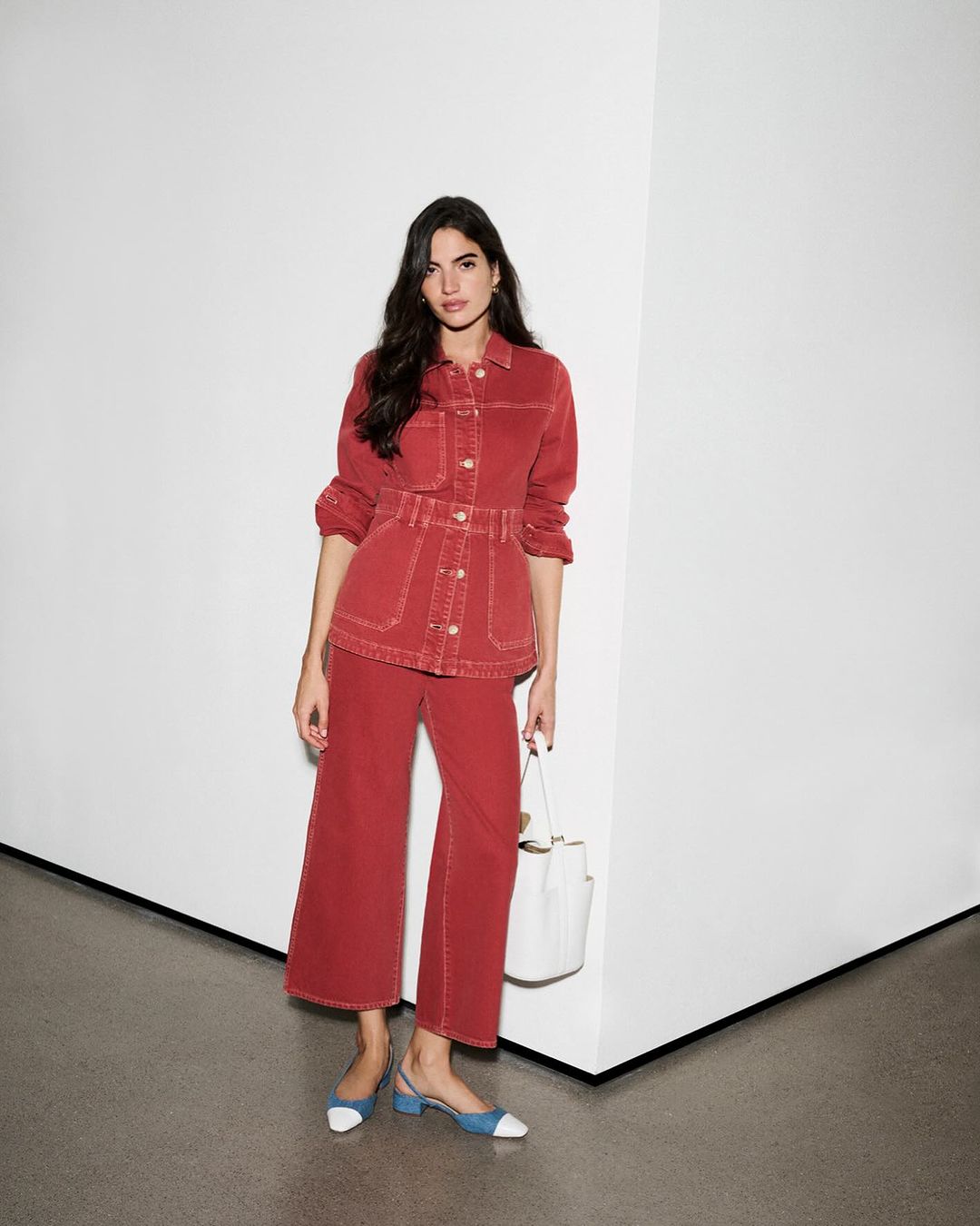 Model in red denim set holding white handbag standing against a white corner wall photographed by Emma Anderson