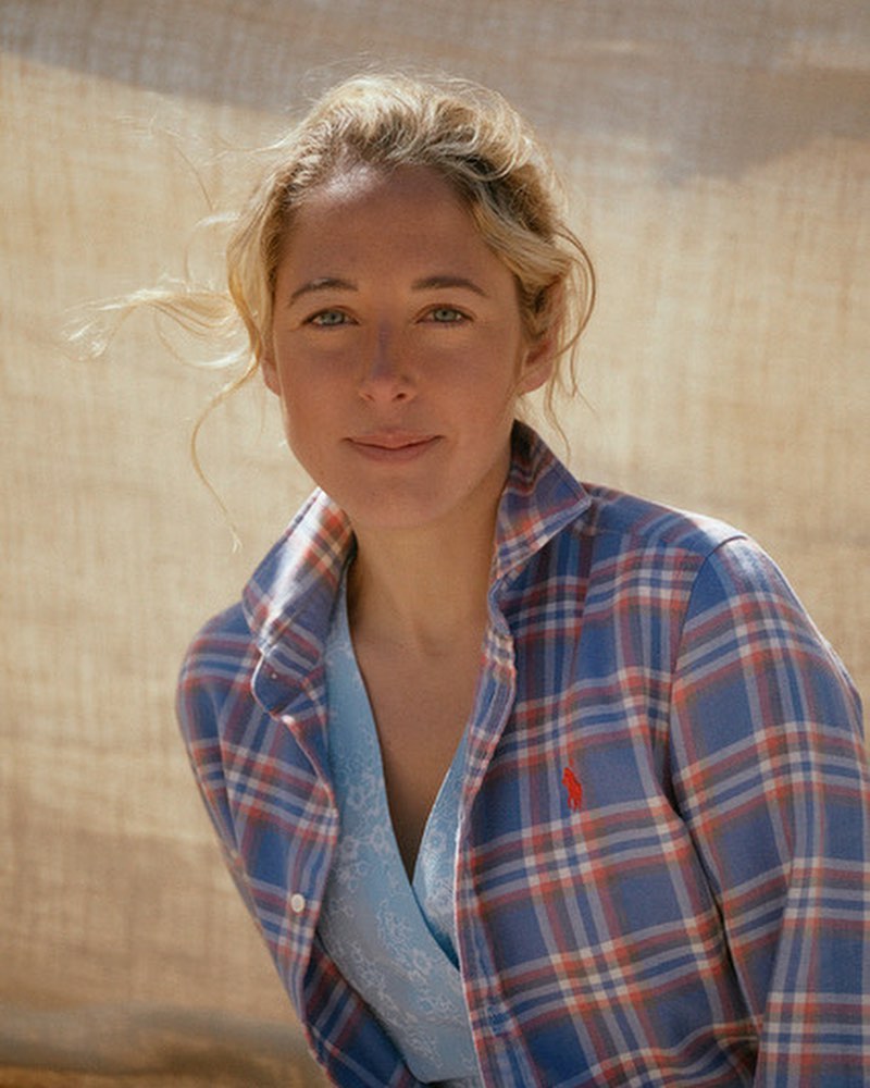 Portrait of a fair-skinned woman with blonde hair smiling in a blue and red plaid shirt, captured with natural lighting.