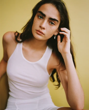 Close-up portrait of a young woman with freckles wearing a white ribbed tank top against a yellow background.
