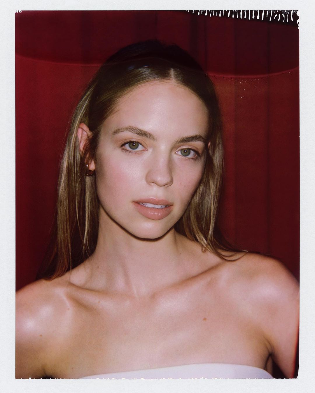 Close-up portrait of a young woman with long brown hair, light skin, and a neutral expression against a blurred red background.