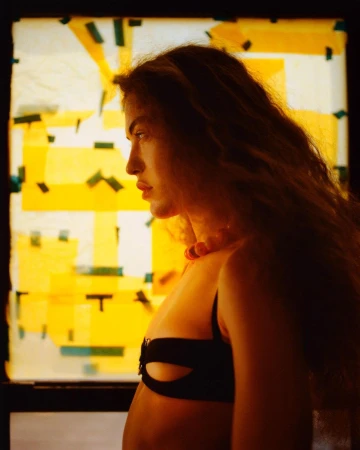Profile portrait of a woman with long wavy hair wearing a black cutout top against a bright yellow and white translucent background.