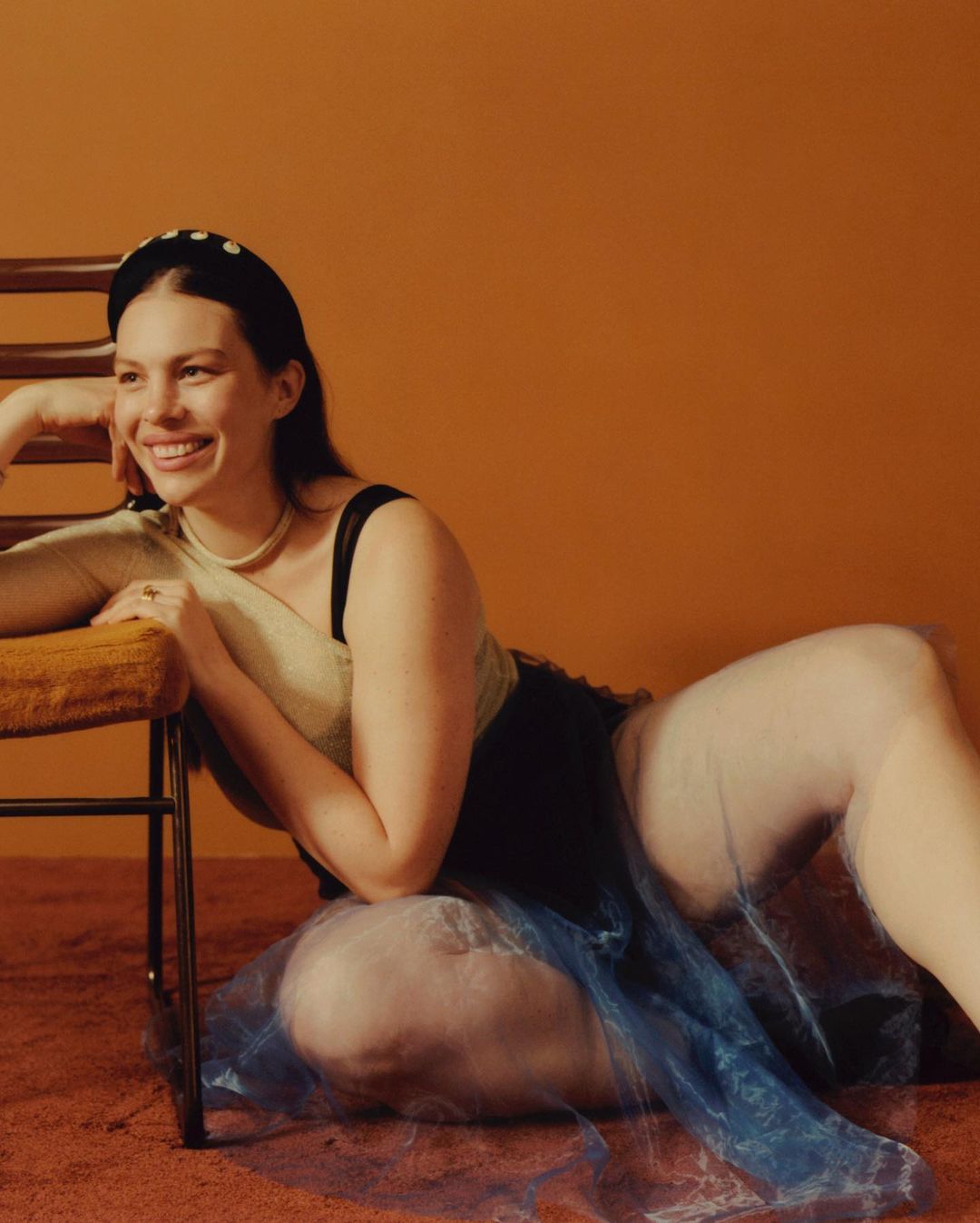 Woman with fair skin and a happy expression is lying on the floor next to a chair against a reddish-brown background.