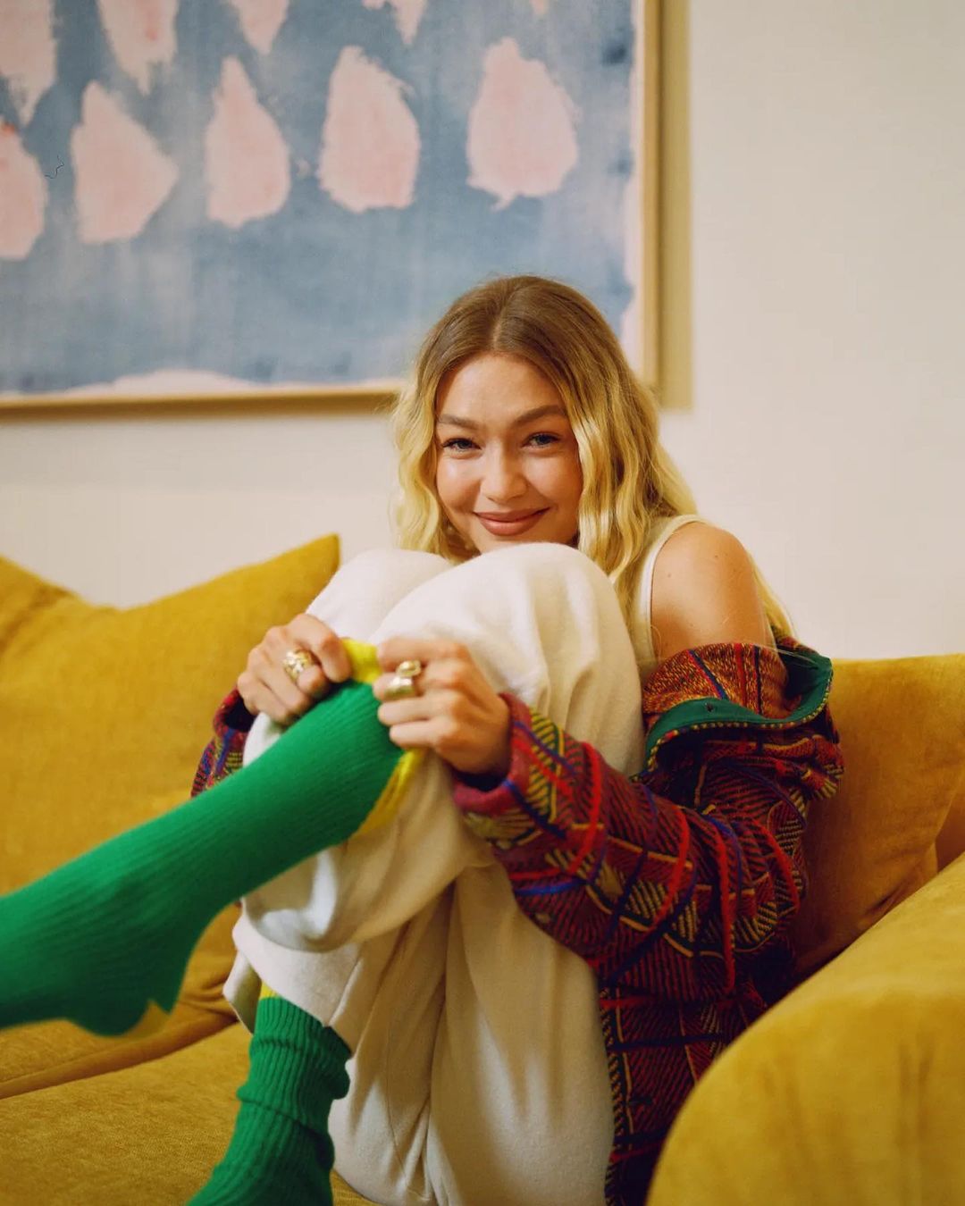 Blonde woman in casual outfit smiles while sitting on a yellow couch in an indoor setting with a colourful painting in the background.