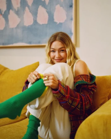Blonde woman in casual outfit smiles while sitting on a yellow couch in an indoor setting with a colourful painting in the background.
