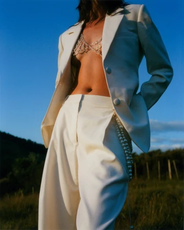 Model wearing a white suit jacket and trousers with a beaded bralette against a bright blue sky and grassy field.
