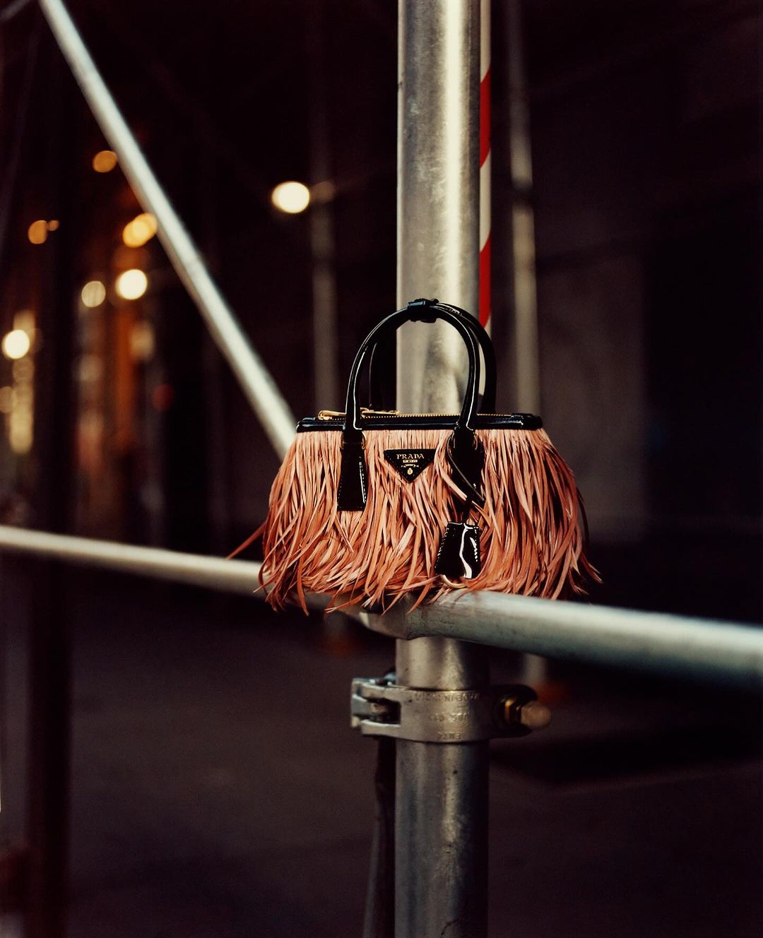 Prada handbag with fringe detail hanging from scaffolding outdoors, set against dark urban background with blurred lights.