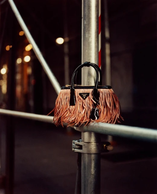 Prada handbag with fringe detail hanging from scaffolding outdoors, set against dark urban background with blurred lights.