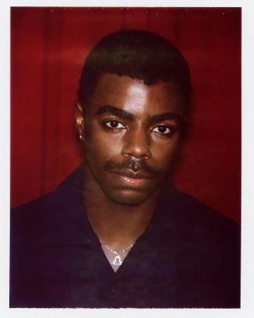 Polaroid portrait of a Black man with a mustache wearing a dark shirt and silver pendant necklace against a red background.