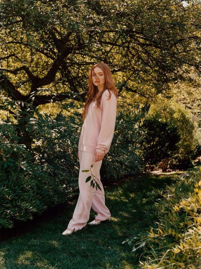 A young woman with red hair stands in a green garden setting, wearing a pink lounge set and holding a leafy stem, outdoors.