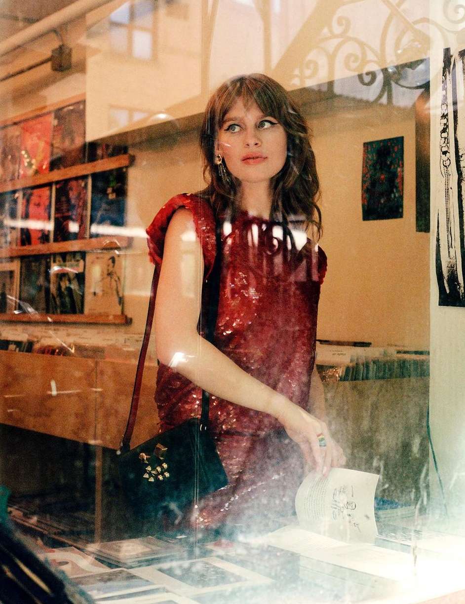 Woman in a red sequin dress standing in front of a shop window, wearing a black purse, posing for a fashion shot.