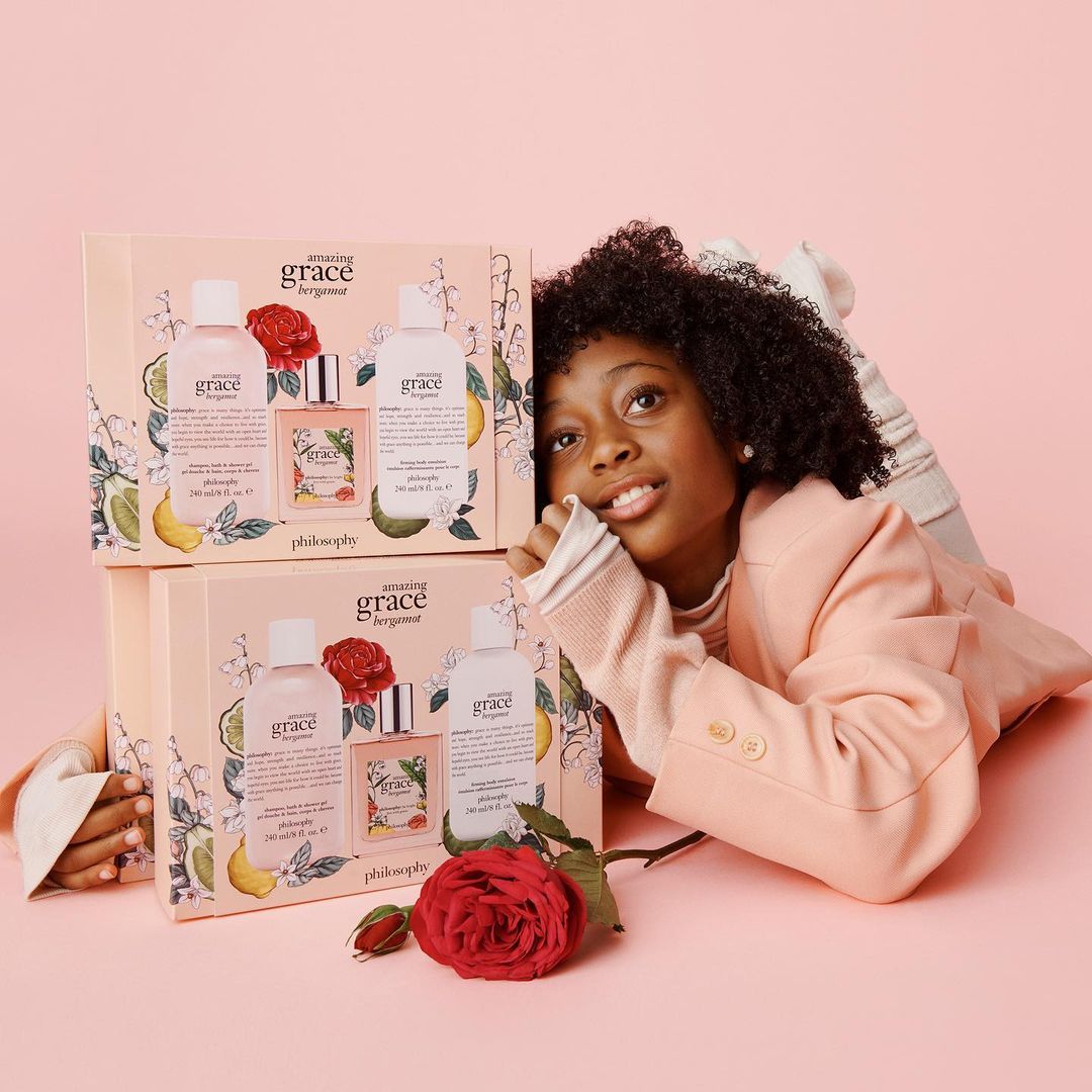 A young African-American woman in a peach blazer poses next to stacked Philosophy Amazing Grace Bergamot gift sets on a peach background.