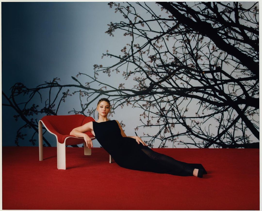An adult female reclines in a red chair on a red surface, wearing a sleeveless black gown, against a backdrop of a white cherry blossom tree.