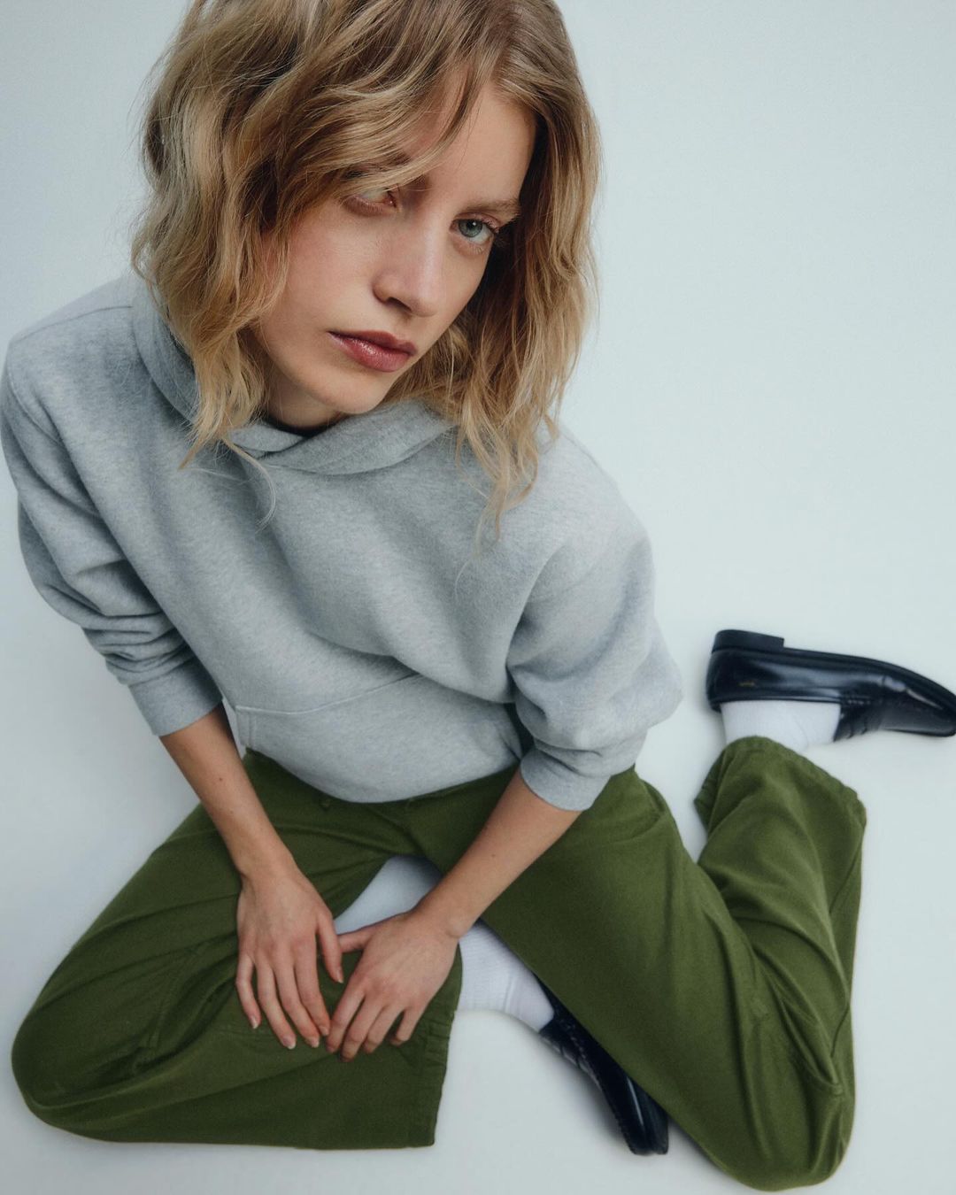 A young woman with blonde hair poses in a grey hoodie and green pants with black loafers, set against a white studio background.