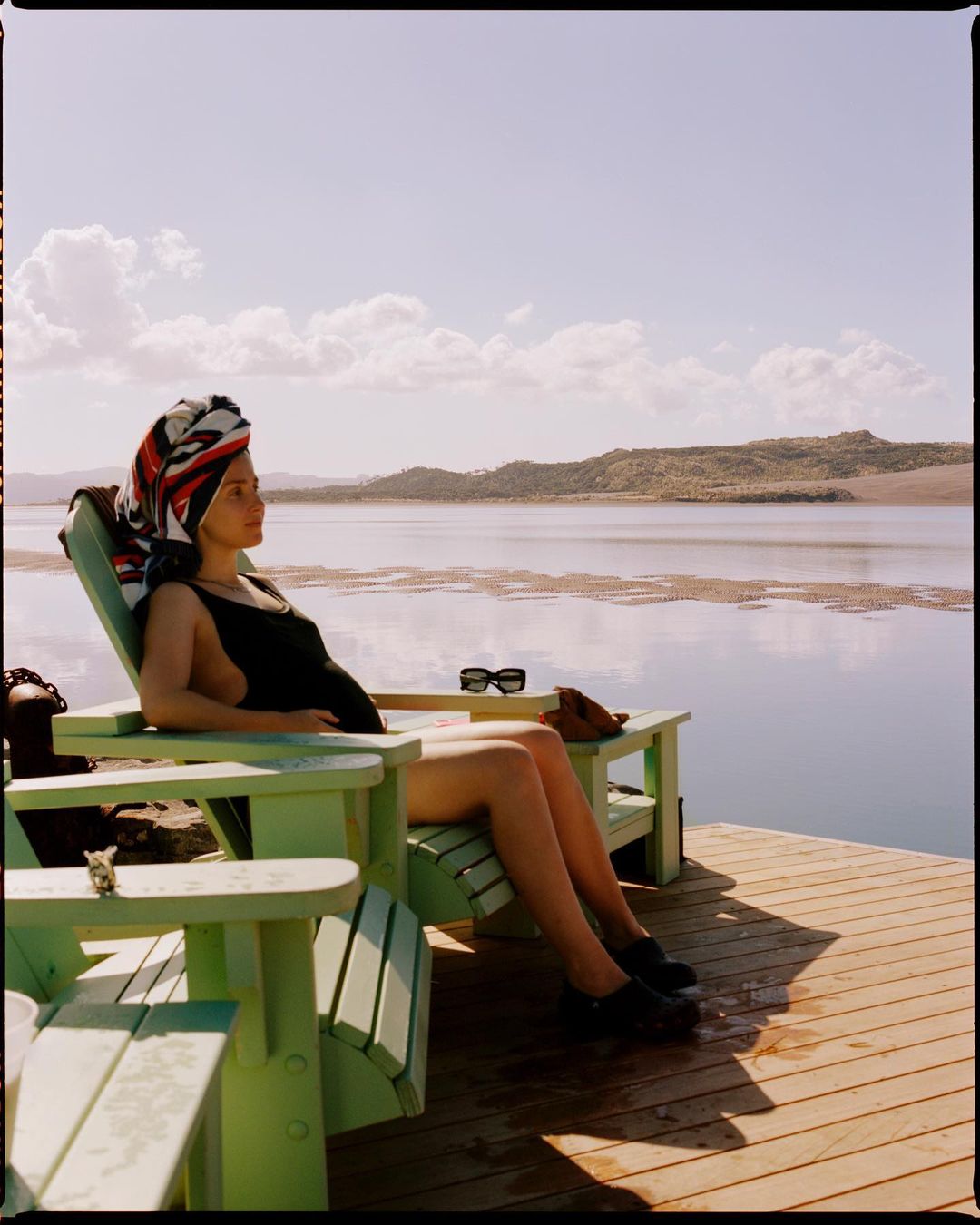 Adult woman in a black swimsuit and headscarf sitting on a green lounge chair by the water with blue sky and clouds in the background.