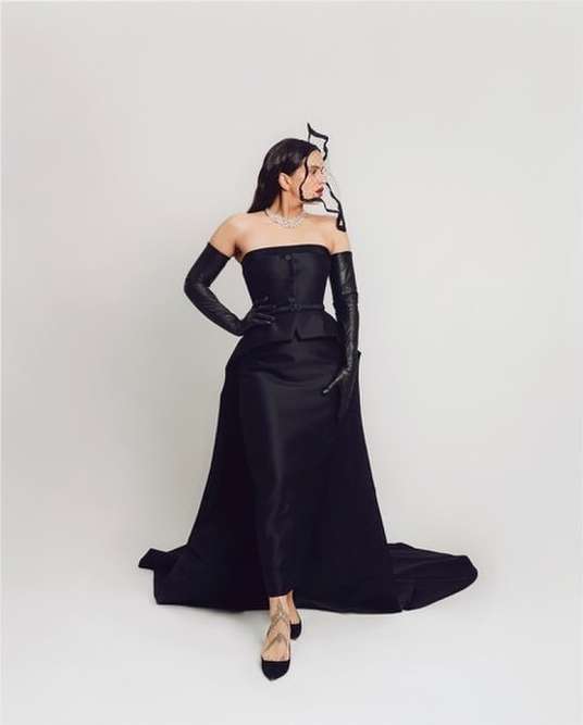 A woman in an elegant black strapless gown with long gloves strikes a pose against a white background, showcasing high fashion.