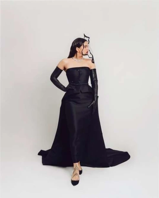 A woman in an elegant black strapless gown with long gloves strikes a pose against a white background, showcasing high fashion.