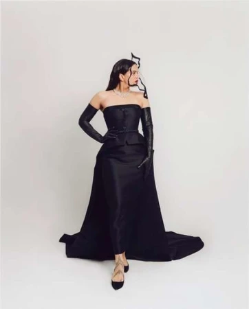 A woman in an elegant black strapless gown with long gloves strikes a pose against a white background, showcasing high fashion.