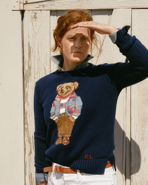 Woman wearing a Polo Bear sweater and white jeans shielding her eyes in the bright sun against a light-colored wooden wall.