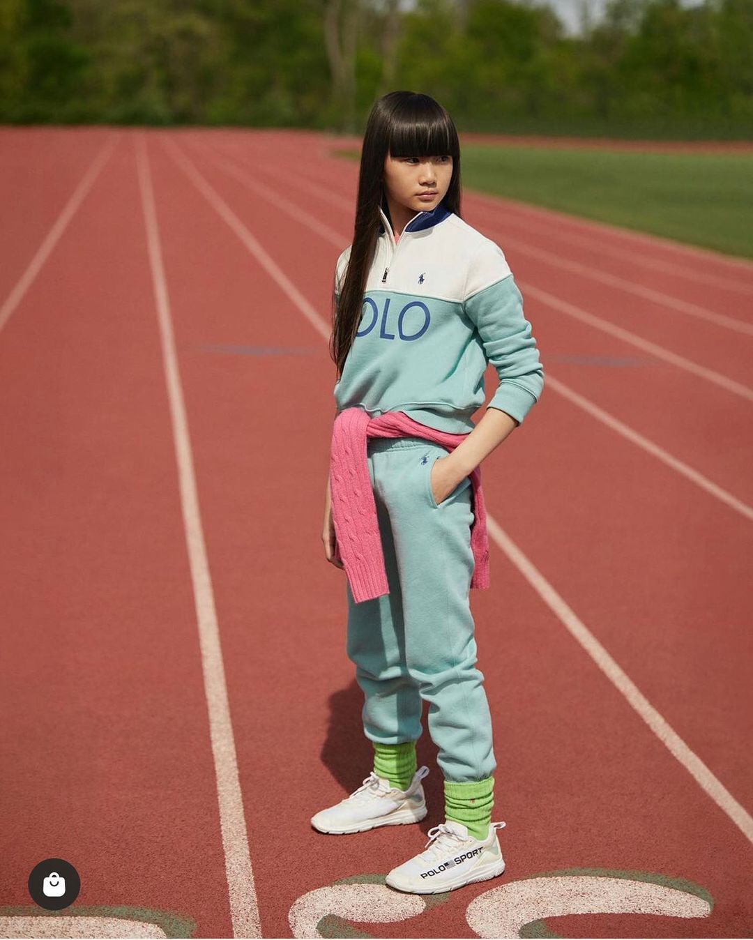 Young girl stands on a red running track wearing athletic clothing and white sneakers with a pink sweater tied around her shoulders.