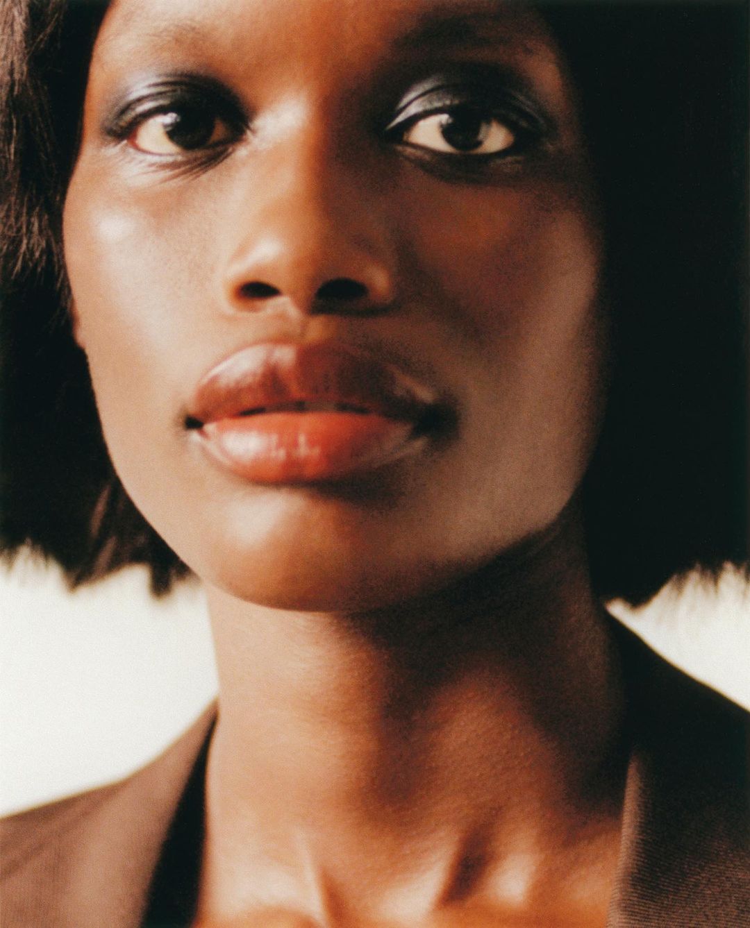 Close-up portrait of a woman with dark hair and glowing brown skin, wearing a brown blazer. Studio lighting creates a soft, focused image.