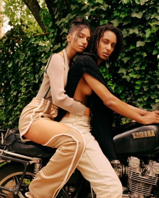 Two models, one male and one female, pose on a black Yamaha motorcycle outdoors with lush green ivy in the background in a medium shot.