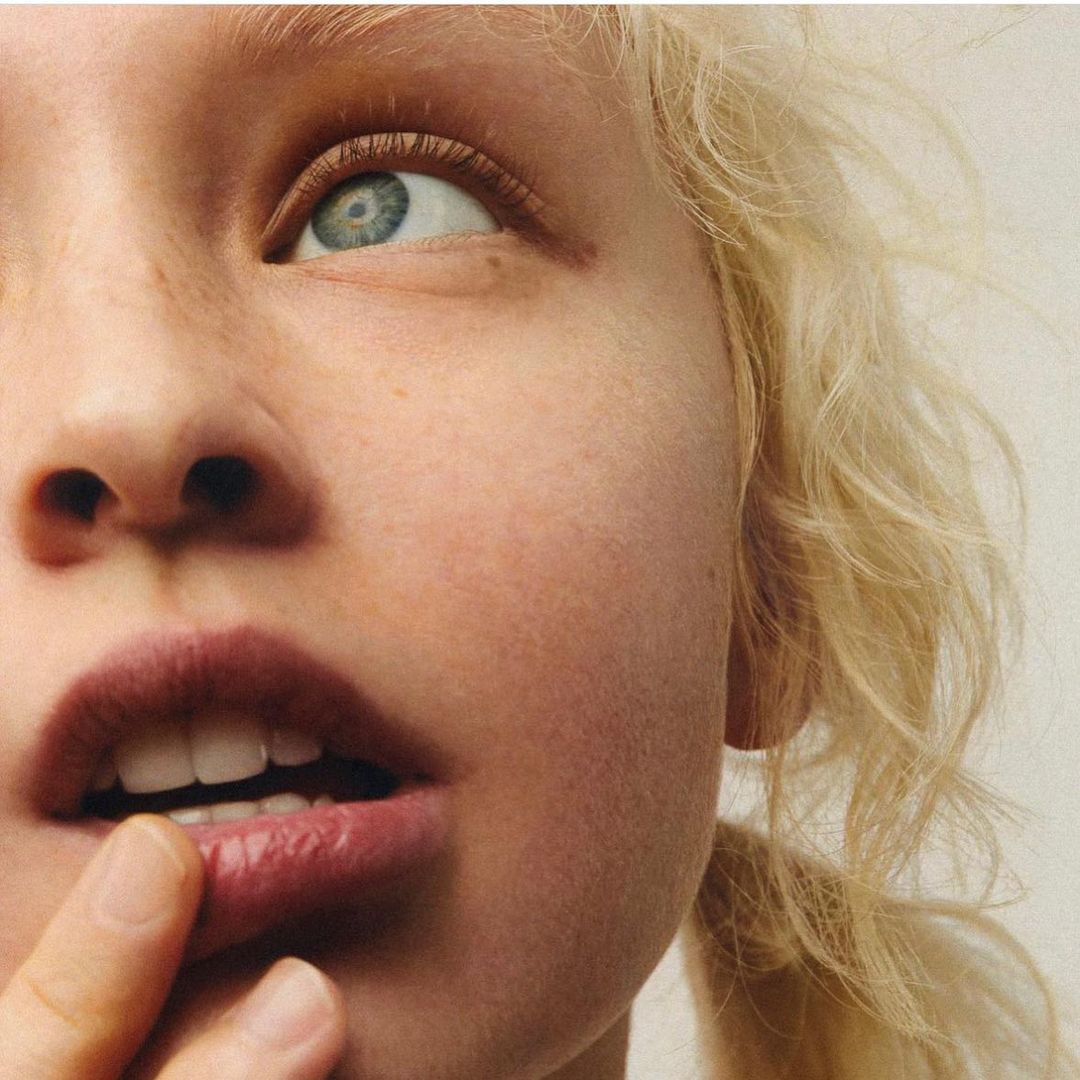 Close-up shot of a blonde adult female with light skin, blue eyes, and a neutral expression, with finger touching the lower lip.