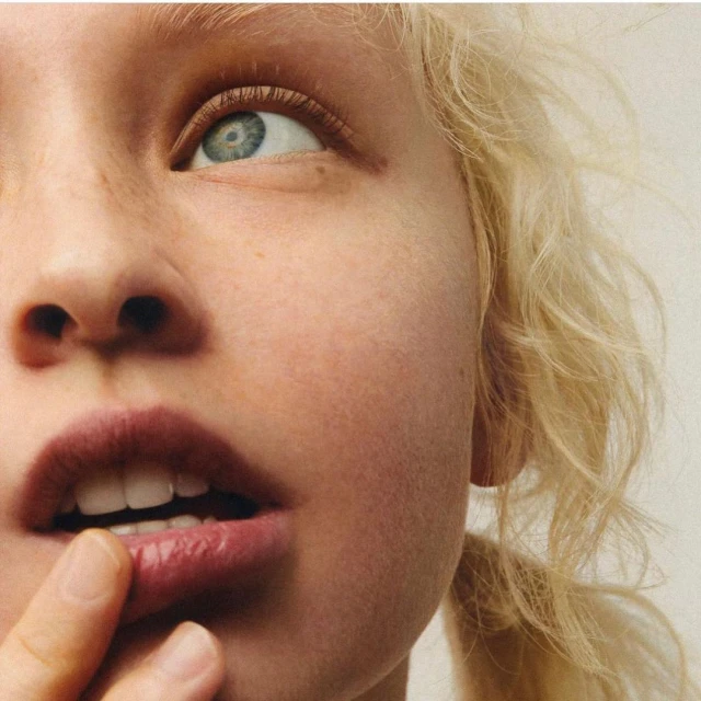 Close-up shot of a blonde adult female with light skin, blue eyes, and a neutral expression, with finger touching the lower lip.