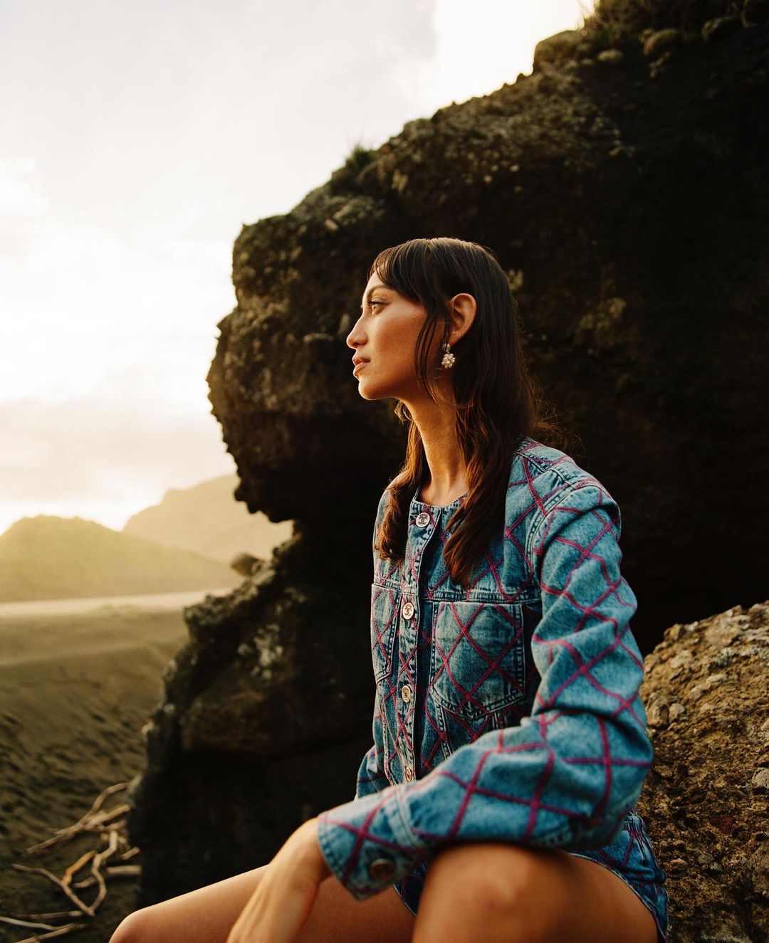 Woman in denim jacket gazes at black sand beach near rocky outcrop during golden hour, capturing a moment of pensive reflection.