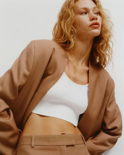 Woman in a brown suit jacket and pants with a white ribbed top, posing in a bright photo studio with soft, diffused lighting.