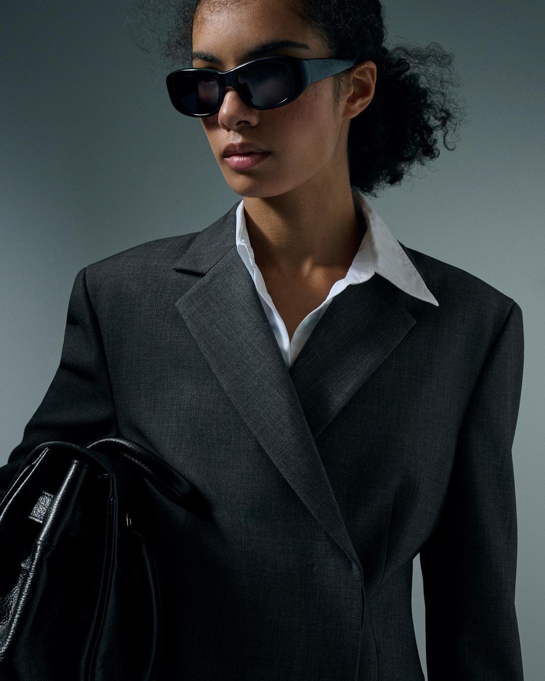Portrait of a woman wearing dark sunglasses, white collared shirt, and charcoal blazer holding a black handbag.