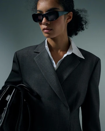 Portrait of a woman wearing dark sunglasses, white collared shirt, and charcoal blazer holding a black handbag.