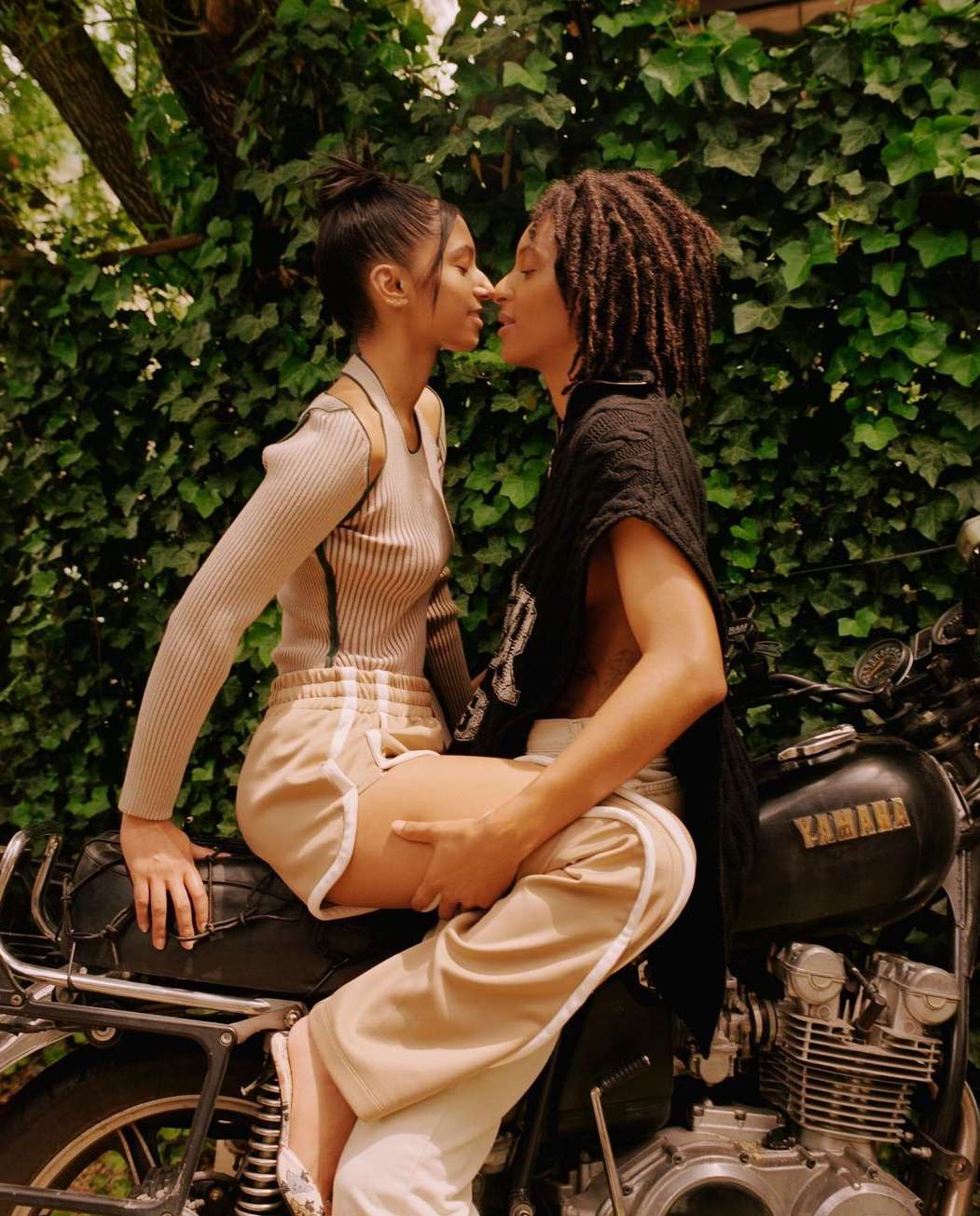 Two adults sit on a motorcycle outdoors, against a leafy backdrop. One person wears a light brown top and shorts, the other wears a black top.