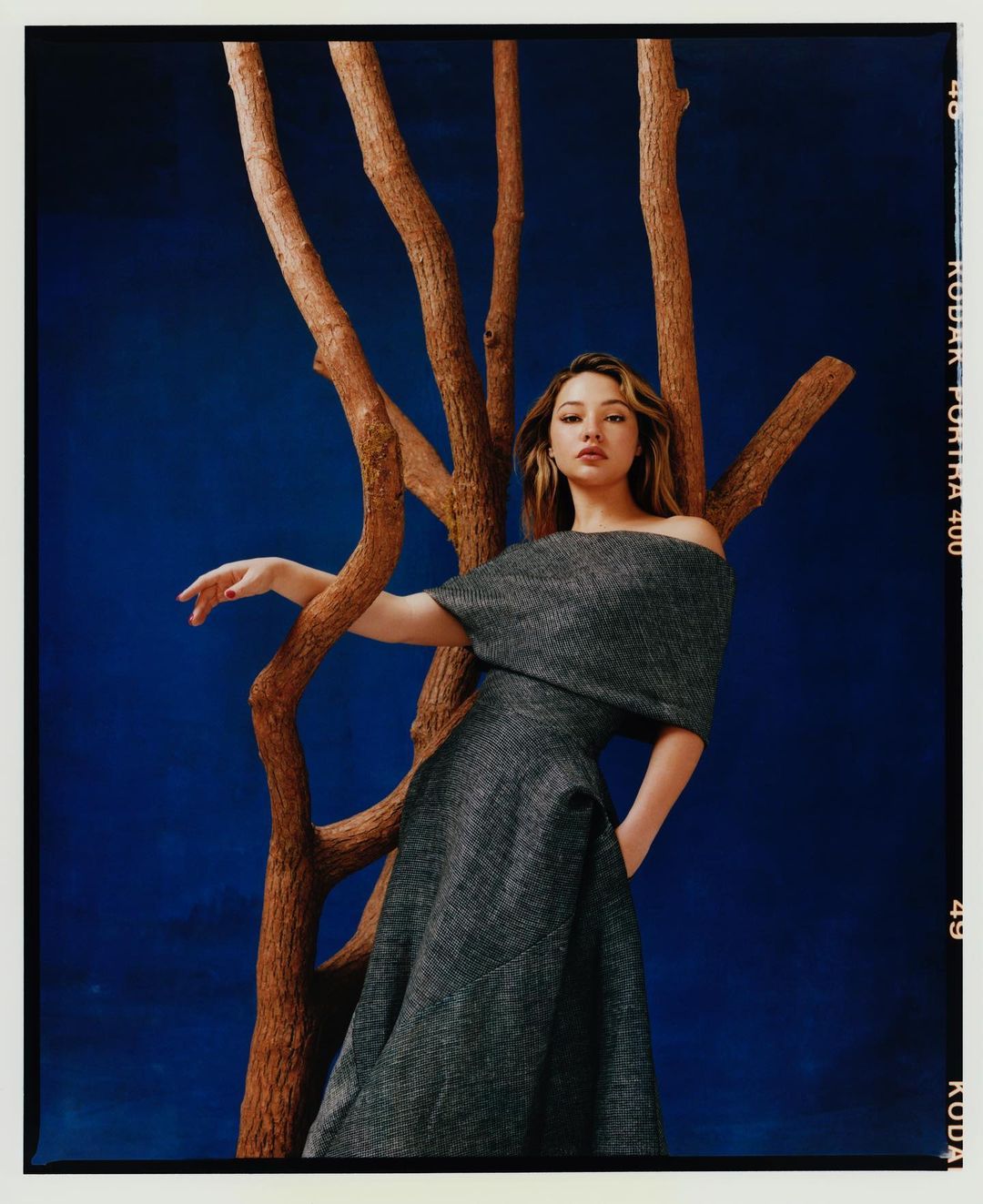 A woman in an off-the-shoulder dress poses with a textured tree branch against a deep blue backdrop in a studio portrait.