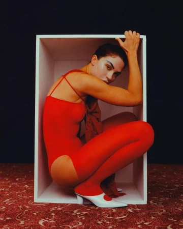 Woman in a red leotard and tights poses inside a white box, striking a crouched pose, with a dark backdrop and patterned floor.