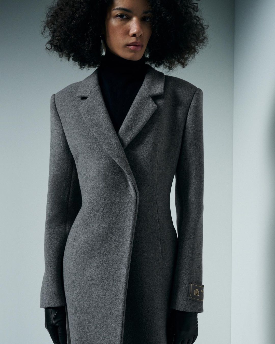 A woman with curly hair poses in a grey wool coat with black gloves, set against a neutral studio backdrop. Fashion editorial shot.