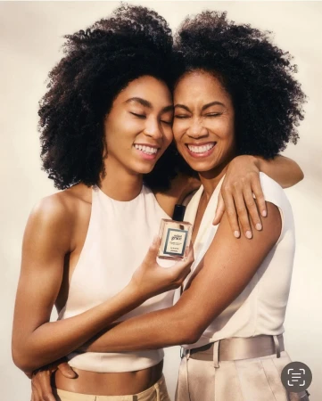 Two smiling Black women embracing while holding a perfume bottle in a studio portrait by Emma Anderson