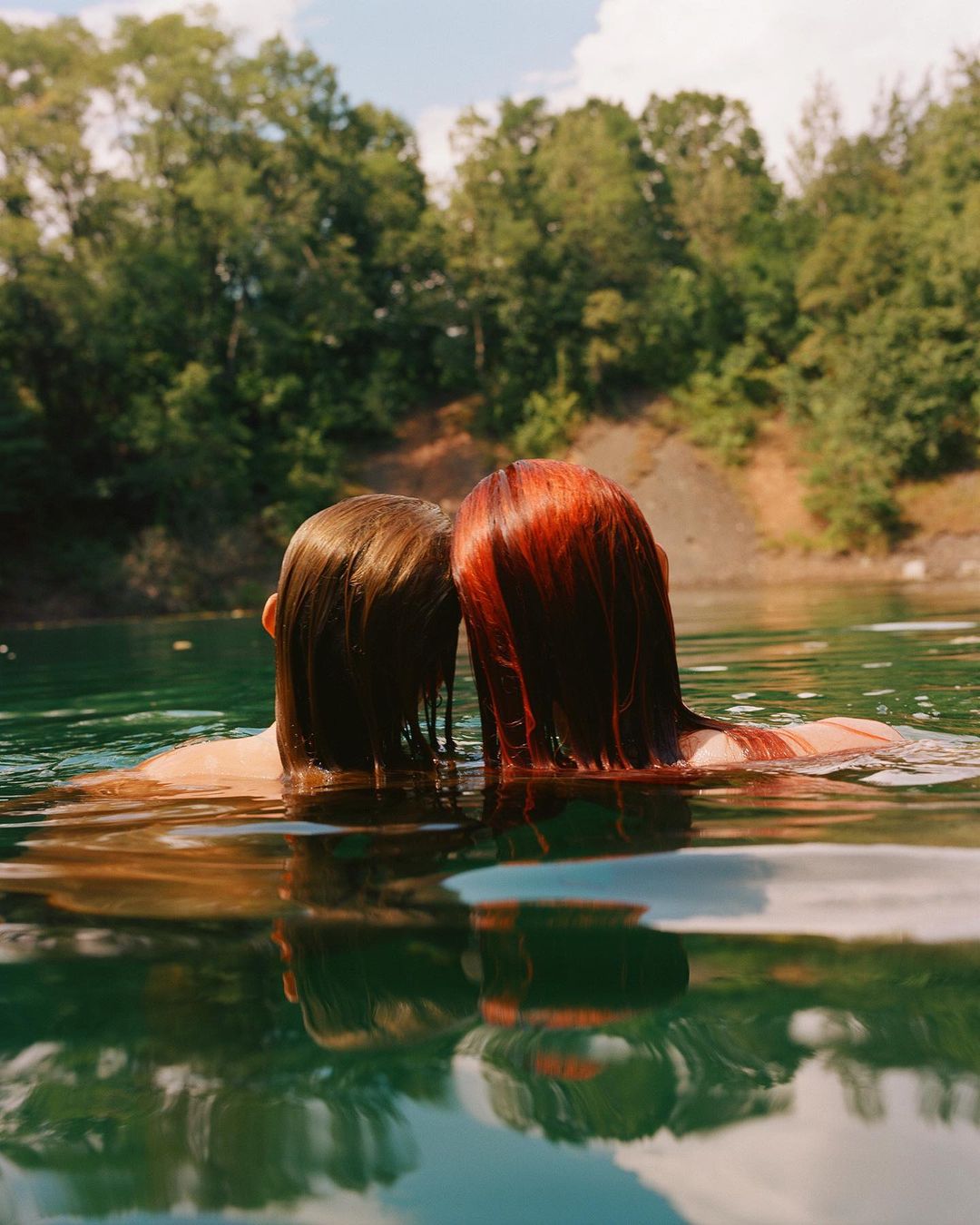 Two people with wet hair submerged in a lake with lush green trees lining the background shoreline