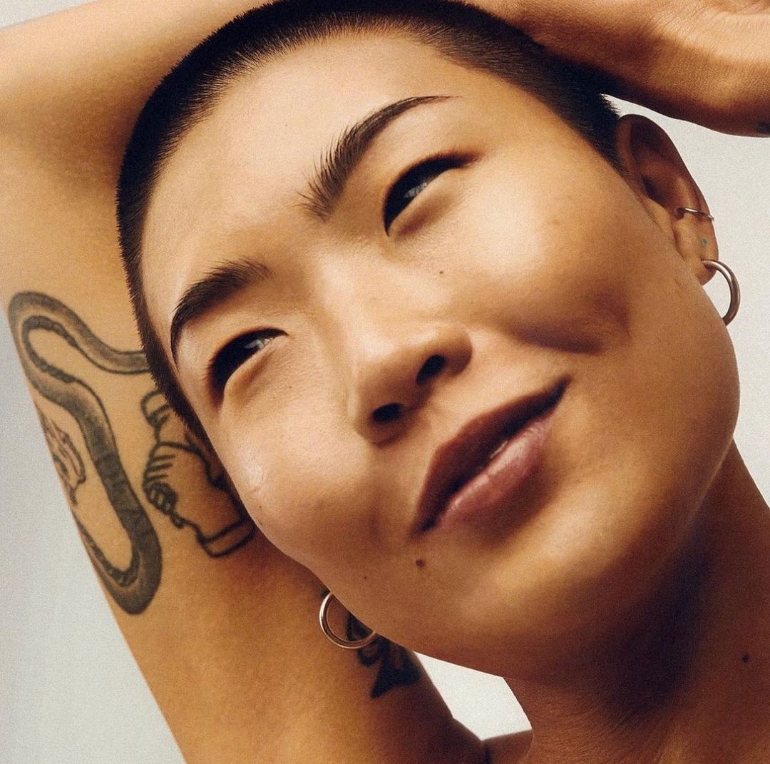 Close-up portrait of a smiling Asian woman with a shaved head and arm tattoo against a light background. Fashionable, with hoop earrings.