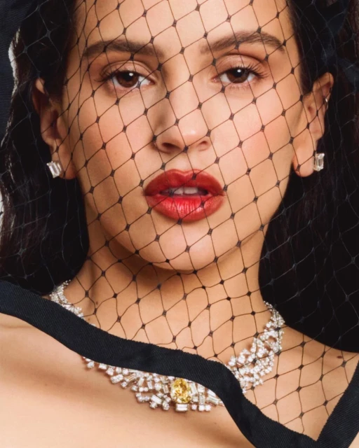 Close-up portrait of a woman with red lipstick wearing a black veil and a diamond necklace against a dark background.