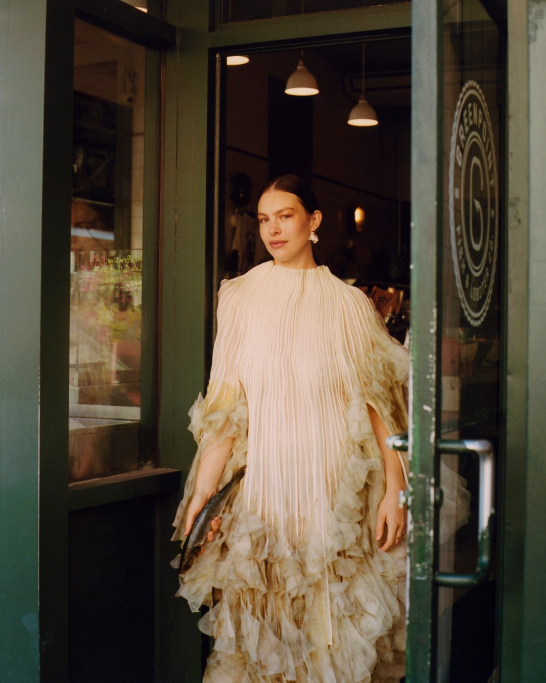 A fashionable woman in an elegant dress is walking through the green door of an establishment with white pendant lights and logo.