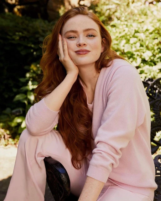 Red-haired woman in a light pink sweater and pants sits outside with her hand to her face, looking directly at the camera.