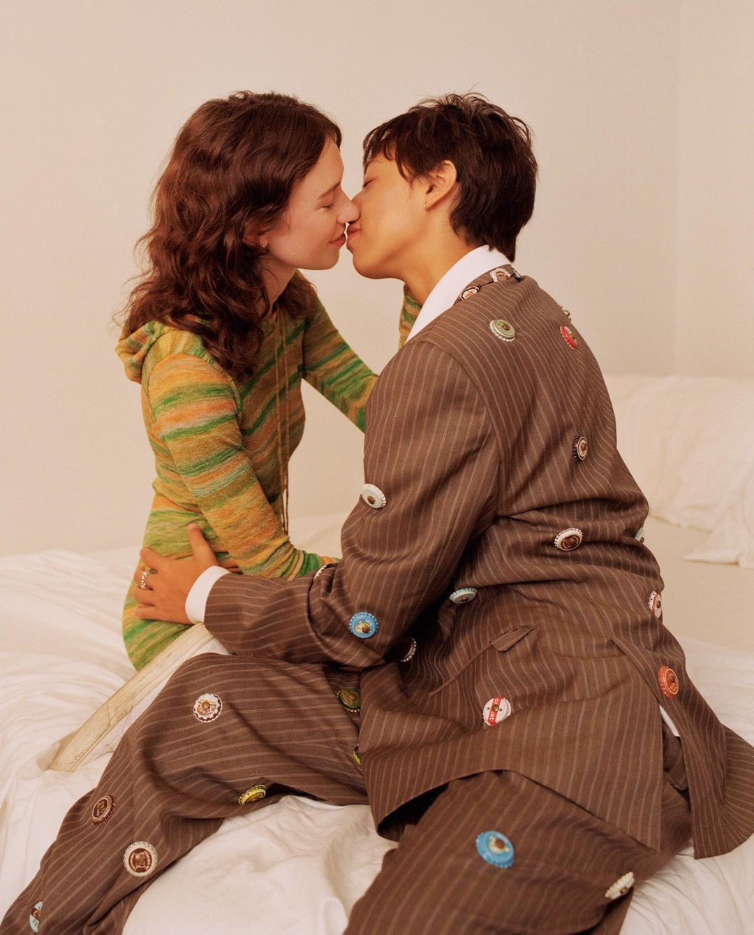 Two young adults are kissing, one with short dark hair wearing a brown pinstripe suit with button embellishments, sitting on a bed.