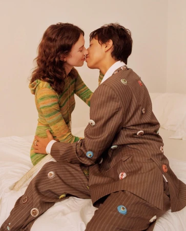 Two young adults are kissing, one with short dark hair wearing a brown pinstripe suit with button embellishments, sitting on a bed.