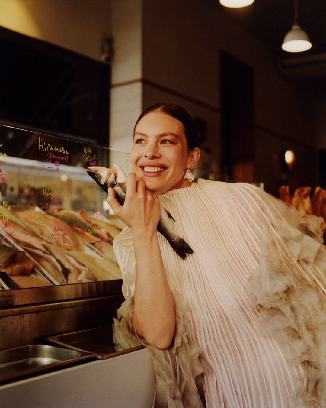 Smiling woman in ruffled dress holds a fish next to a seafood display, indoor setting, warm lighting, commercial environment.